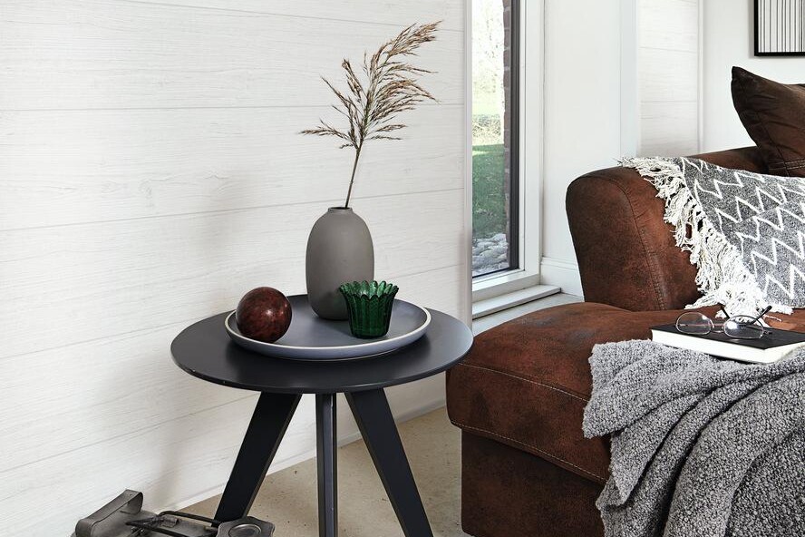 Dekorpaneele als zeitlose Wandverkleidung im Wohnzimmer Weiße Wandpaneele mit Struktur, kleiner Beistelltisch mit Deko und braunes Sofa